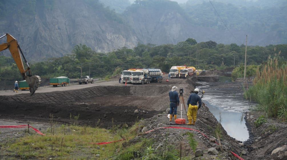 Ingenieure vermessen das Gelände zwischen einem mit Rohöl gefüllten Erdteich rechts und der links sichtbaren Schaufel eines Baggers, der gerade eine weitere Erdgrube aushebt. Im Hintergrund stehen Tanklaster vor der Bergkulisse