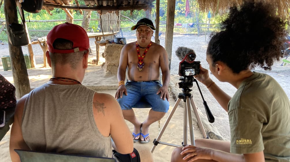 Eine Journalistin und Journalist im Vordergrund nehmen mit einer Kamera auf Stativ ein Interview mit einem indigenen Führer (im Hintergrund) auf