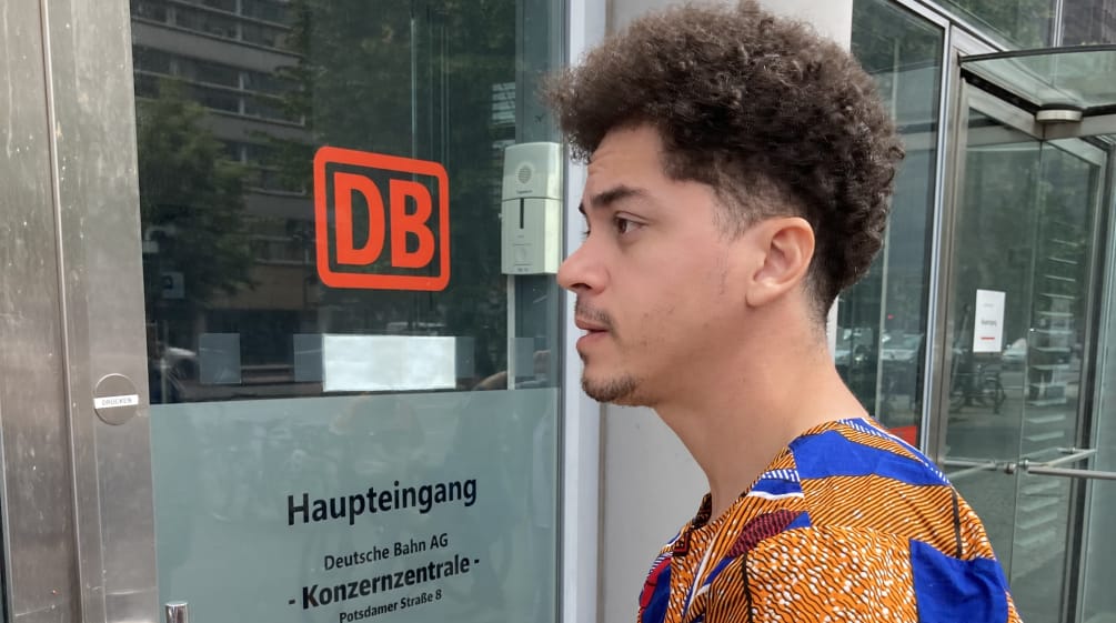 Mikaell von der brasilianischen Organisation Justiça nos Trilhos vor der DB-Zentrale in Berlin Ein junger Mann steht mit einem Briefumschlag vor einer Glastür mit der Aufschrift DB Haupteingang Konzernzentrale