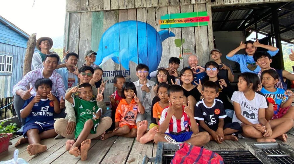 Eine große Gruppe Kinder und Jugendliche sitzen auf der Terrasse eines Holzhauses und zeigen mit den Fingern Begeisterung