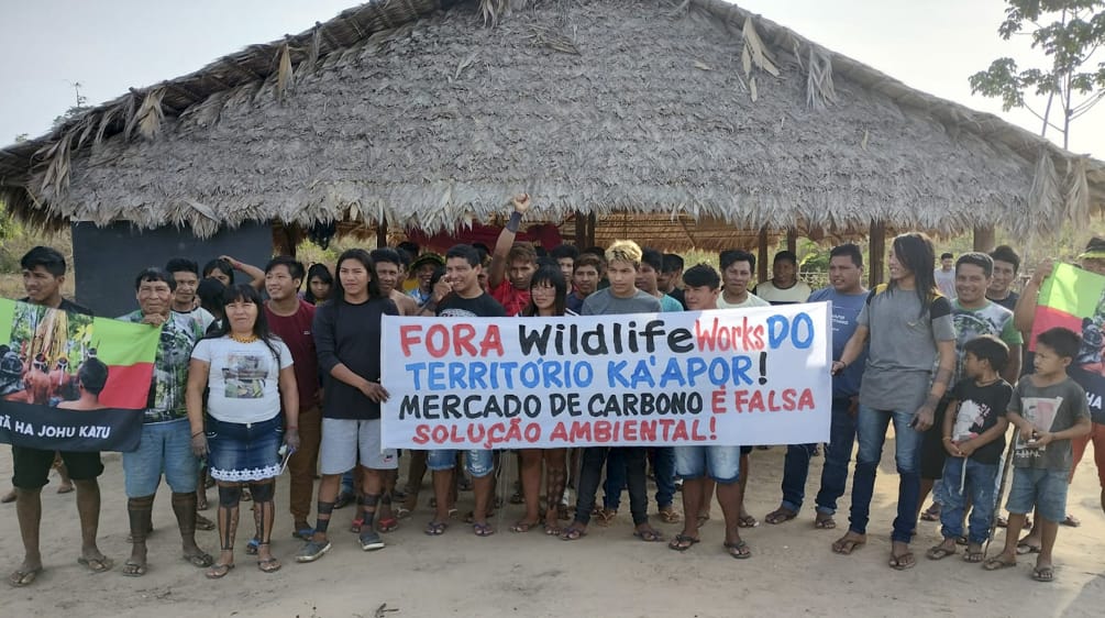Eine Gruppe von Indigenen protestiert mit einem Banner vor einer mit Palmenwedeln überdachten Hütte. Aufschrift des Banners: "Wildlife Works raus aus dem Territorium der Ka'apor. Der Kohlenstoffhandel ist eine falsche Umweltlösung"