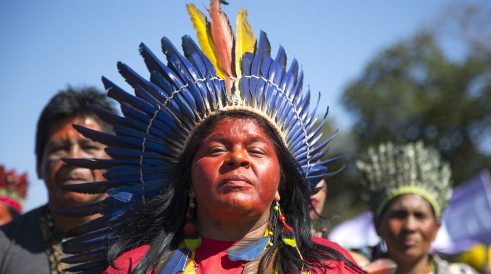 Eine mit einer Federkrone geschmückte indigene Frau vor zwei weiteren indigenen Personen