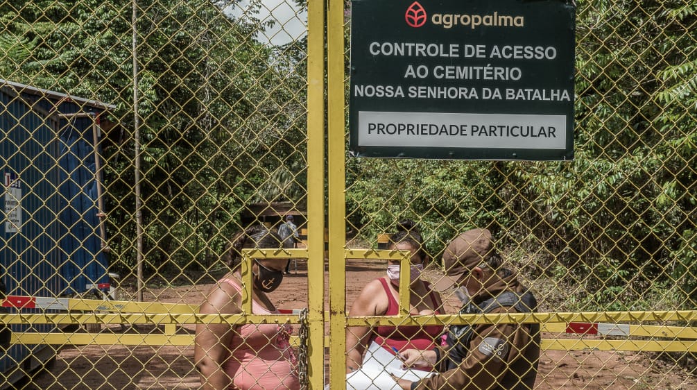 Selbst um auf ihren Friedhof zu gehen, muss die lokale Bevölkerung Zäune und Wachposten von Agropalma überwinden. Zwei Frauen hinter einem hohen Gittertor werden von einem Wachmann kontrolliert. Aufschrift auf Firmenschild: Agropalma. Kontrolle des Zugangs zum Friedhof Nossa Senhora da Batalha. Privatbesitz