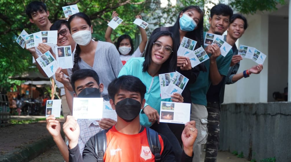 Eine Gruppe Jugendlicher mit Masken hält bunte Postkarten mit Bildern von der Insel hoch