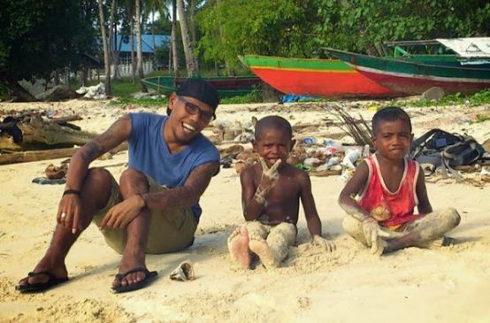 Jopi Peranginangin sitzt mit zwei kleinen Jungen am Strand und lacht in die Kamera