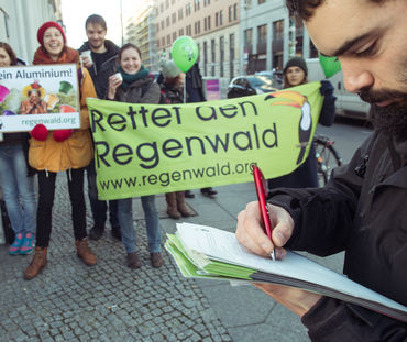 Ein Mann unterschreibt eine Petition. Im Hintergrund Aktivisten mit Banner, Plakaten und Kaffeetassen