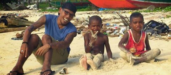 Jopi Peranginangin sitzt mit zwei kleinen Jungen am Strand und lacht in die Kamera