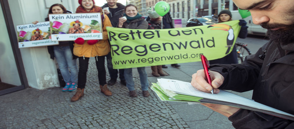 Nespresso soll aus dem schädlichen Kapselgeschäft aussteigen. Dafür sammeln wir weiter Unterschriften. Ein Mann unterschreibt eine Petition. Im Hintergrund Aktivisten mit Banner, Plakaten und Kaffeetassen