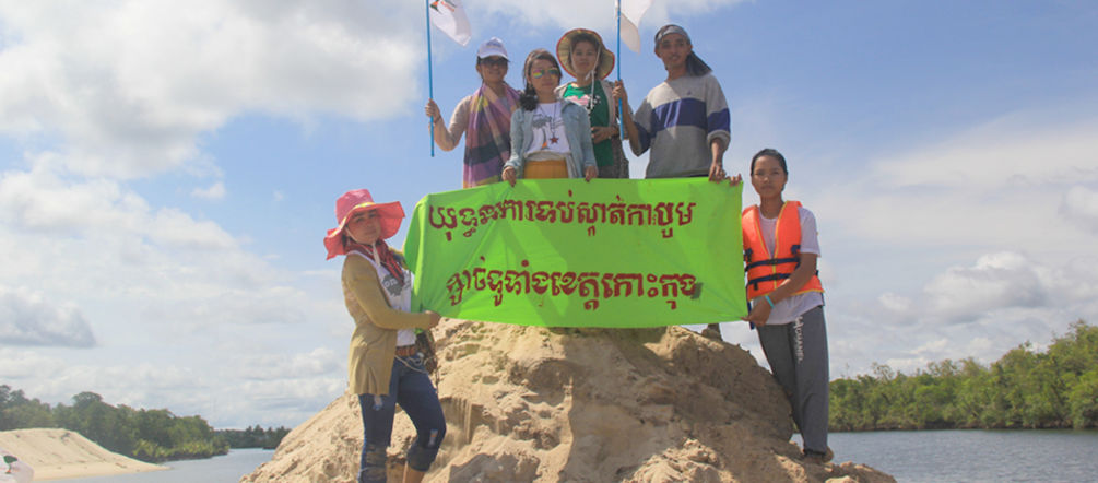 „Stoppt den Sandabbau in der Provinz Koh Kong“, fordern die Umweltschützer von Mother Nature Umweltschützer stehen auf einem aufgeschütteten Sandhügel und halten ein Protestbanner hoch: Kampagne gegen Sandabbau in der ganzen Koh Kong-Provinz