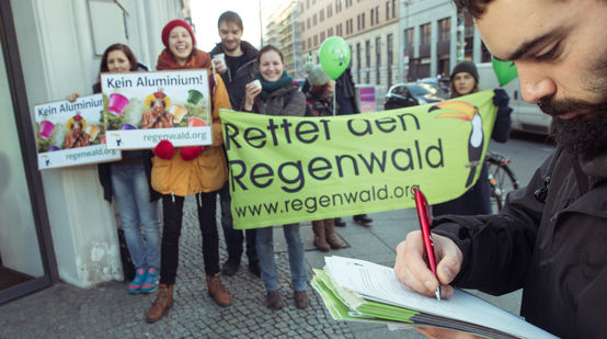 Ein Mann unterschreibt eine Petition. Im Hintergrund Aktivisten mit Banner, Plakaten und Kaffeetassen