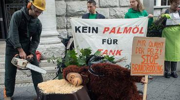 Aktivisten demonstrieren vor dem Gebäude der GIZ in Berlin. Auf einen Baustumpf hat ein Demonstrant im Affenkostüm seinen Kopf gelegt. Dahinter steht ein Mann mit einer Kettensäge. Plakate gegen Palmöl werden hochgehalten.