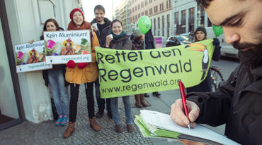 Ein Mann unterschreibt eine Petition. Im Hintergrund Aktivisten mit Banner, Plakaten und Kaffeetassen