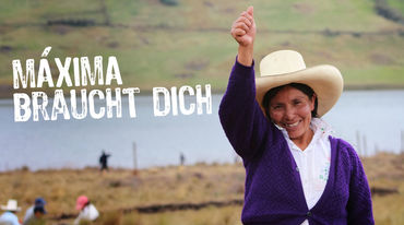 Máxima Acuña erhebt lächelnd ihre rechte Hand. Im Hintergrund sind weitere Bauern und die blaue Lagune zu sehen.
