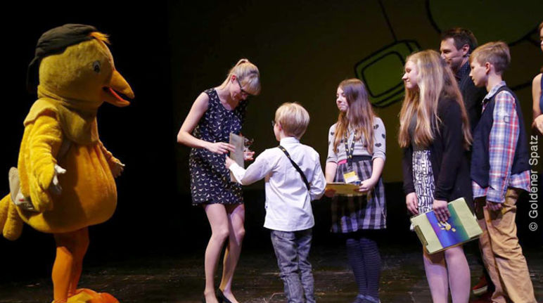 Im Beisein des Festival-Maskottchens nimmt Kathrin Heidtmann den Preis der Kinder-Jury entgegen 5 Kinder überreichen einer Mitarbeiterin von Rettet den Regenwald den Goldenen Spatzen auf der Bühne