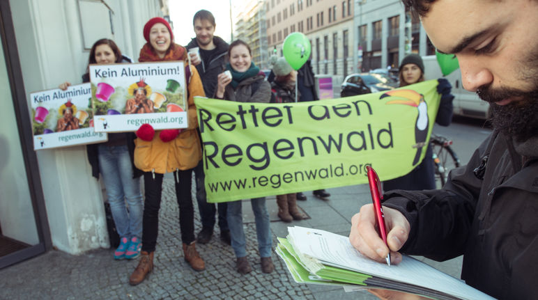 Nespresso soll aus dem schädlichen Kapselgeschäft aussteigen. Dafür sammeln wir weiter Unterschriften. Ein Mann unterschreibt eine Petition. Im Hintergrund Aktivisten mit Banner, Plakaten und Kaffeetassen